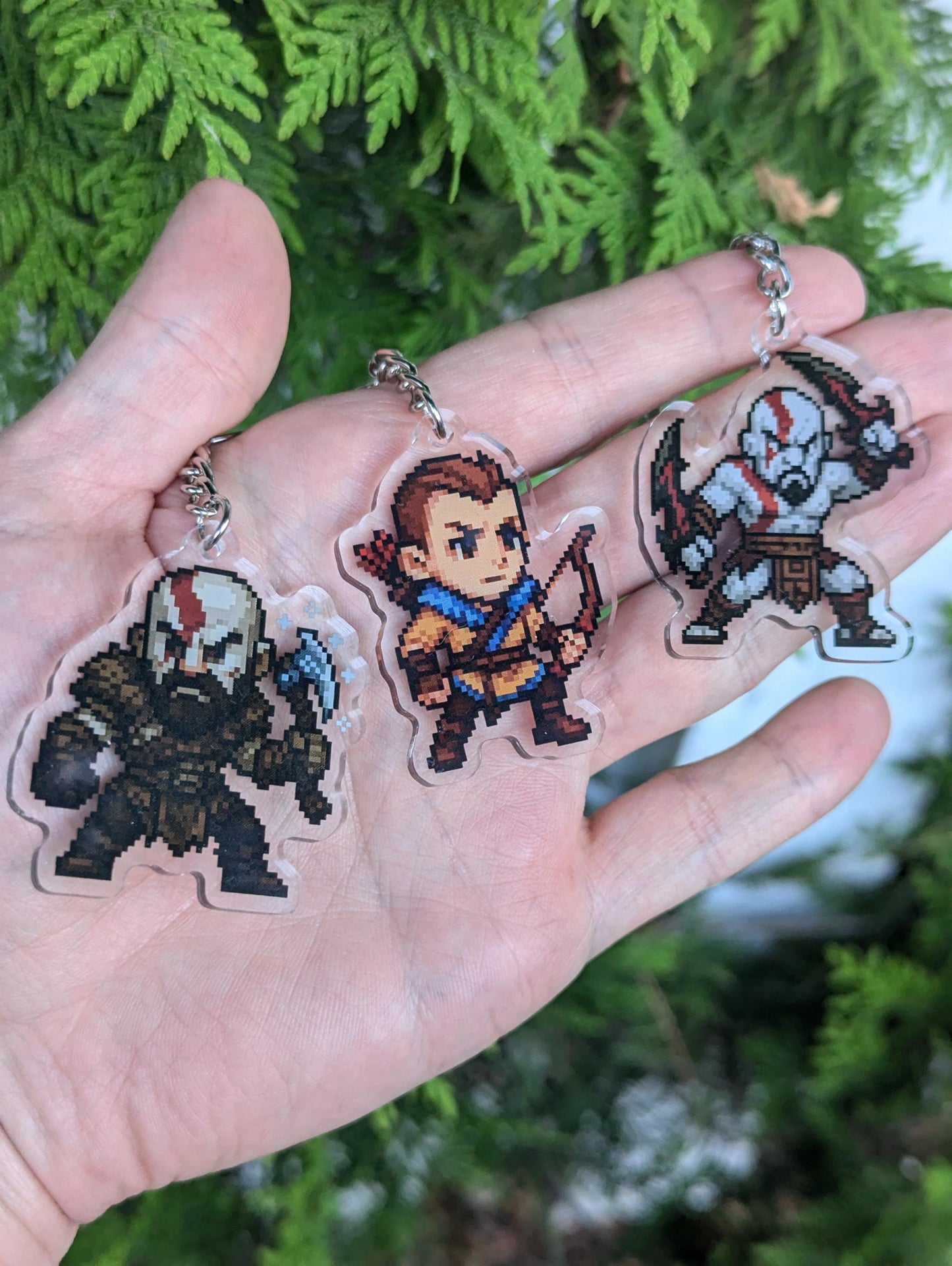 Hand holding 3 pixelized keychains. One is inspired by Kratos from the Norse God of Wars, holding his Leviathon axe while wearing brown armor. His son atreus is by his side wearing a yellow and blue garb seen in the sequel, holding his bow. The 3rd keychain features Kratos from his Greek era, shirtless and holding the Blades of Chaos.