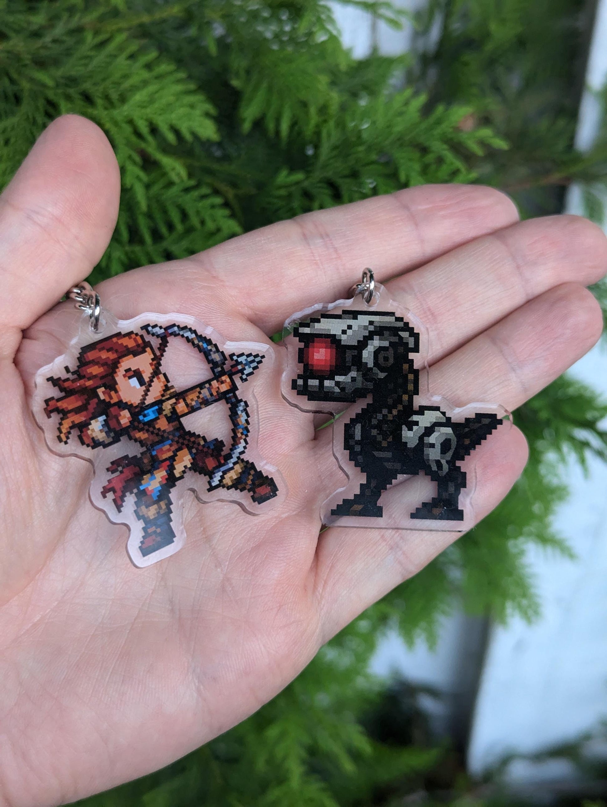 Hand holding acrylic keychains showcasing fan art of Aloy and a watcher from Horizon Zero Dawn & Forbidden West video games. Designs are pixel variants. Aloy features her ginger orange hair, wearing her colorful armor and drawing her bow ready to fire. The watcher is grey and standing in battle stance with a red eye.